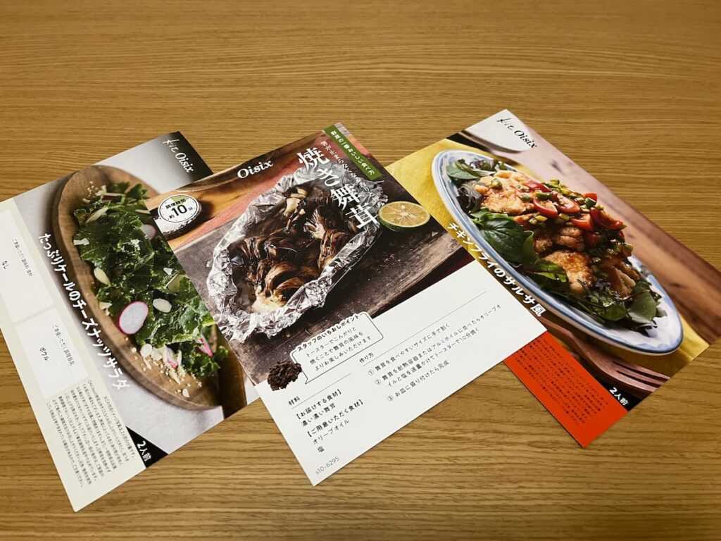 Oisixミールキットのレシピカードを木のテーブルに3枚並べた写真|お試しセットの内容イメージ