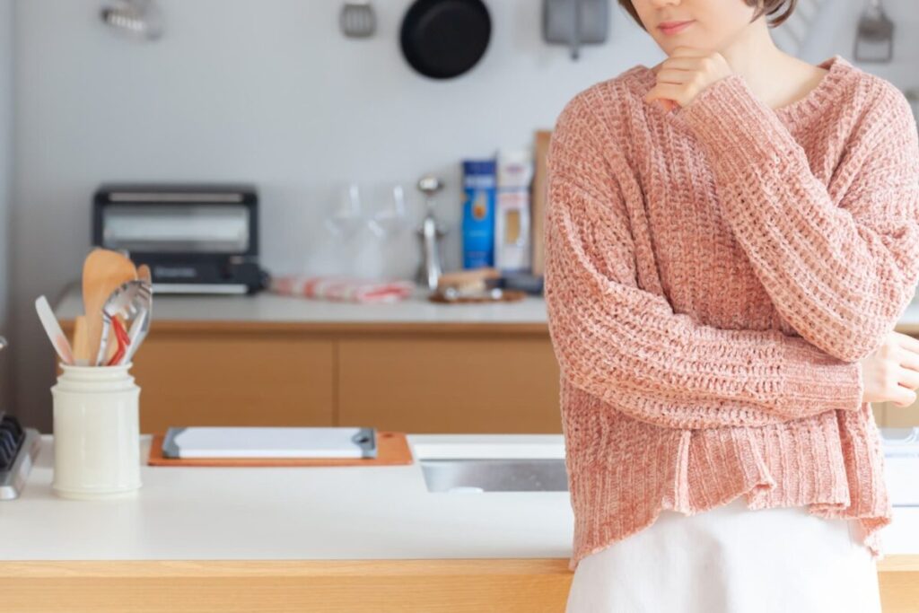 一人暮らしの女性がキッチンで料理に悩んでいる様子の写真|自炊の負担をイメージ